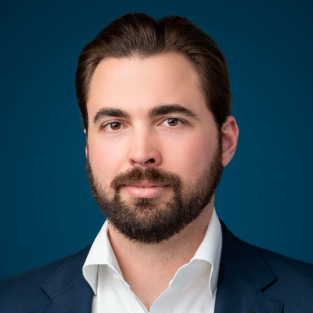 A professional headshot of a man with dark hair and a beard, wearing a white collared shirt and a dark blue blazer, set against a solid dark teal background.