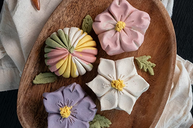 Four intricately sculpted floral wagashi (Japanese confections) in pink, white, purple, and multicolored dough, arranged on a wooden plate with green edible leaves.